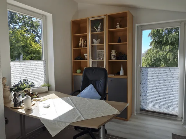 Schreibtisch mit Bürostuhl zwischen zwei Fenstern mit Blick ins Grüne